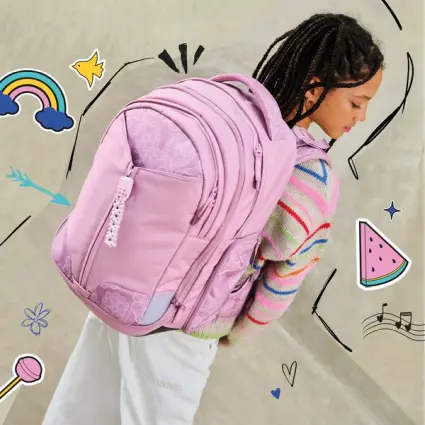 Ein Mädchen mit gelber Mütze und grün-schwarzem satch pack Rucksack steht vor einem Scheinwerfer und dreht sich um zur Kamera.