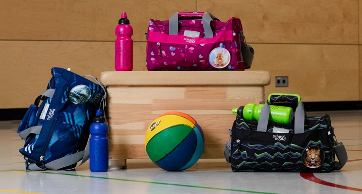 Auf einem Turnhallenboden stehen 3 bunte Sporttaschen und 2 Trinkflaschen von School-Mood