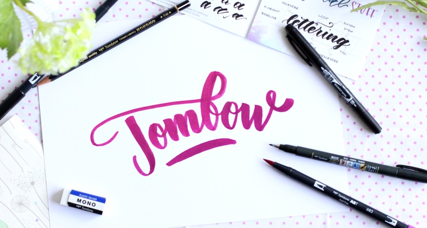 Handlettering mit „Tombow“ in Pink, umgeben von Tombow-Brush Pens und Radierer..
