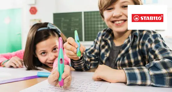 Zwei Kinder sitzen an einer Schulbank und halten Schreiblernfüller von STABILO in die Kamera.