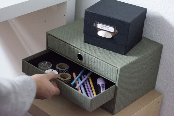 Auf einem Schrank stehen eine salbeifarbene Schubladenbox mit drei Schubladen und eine schwarze Fotobox der S.O.H.O.-Kollektion von Rössler. Die mittlere Schublade wird von einer Hand geöffnet. Darin befinden sich verschiedene Stifte und Washitape von Rössler.