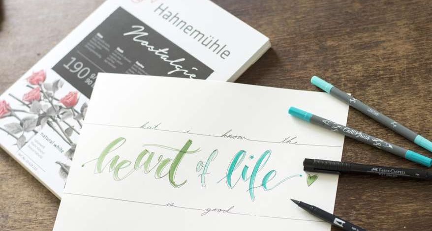 Auf einem Holztisch liegt ein Block mit Zeichenpapier der Marke Hahnemühle. Auf einem Blatt ist in grün und blau gelettert "heart of life" 