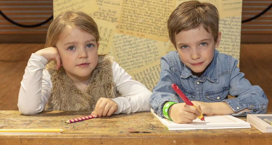 Zwei Kinder, ein Junge und ein Mädchen im Grundschulalter, sitzen an einer Schulbank. Vor ihnen liegt ein aufgeschlagenes Heft, sie haben Stifte in der Hand und blicken in die Kamera.