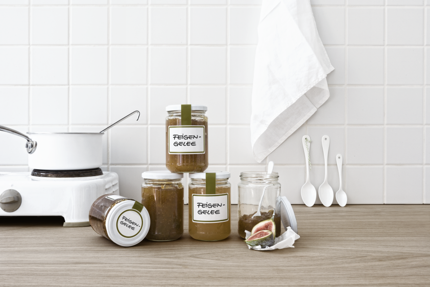 Auf einer Küchenanrichte mit hellem Holz stehen 5 Gläser. Drei davon sind mit einem Etikett der Marke Avery Zweckform beklebt und beschriftet. Links daneben steht ein Topf auf einer Kochplatte, im Hintergrund hängt an der gefliesten Küchenwand ein Abtrockentuch.