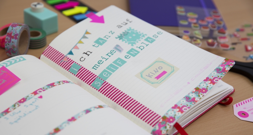 Ein aufgeschlages Notizbuch von Leuchtturm1917 ist mit Maskingtape und einem Spruch bunt verziert. Drunherum liegen Rollen Maskingtape und Sticker.