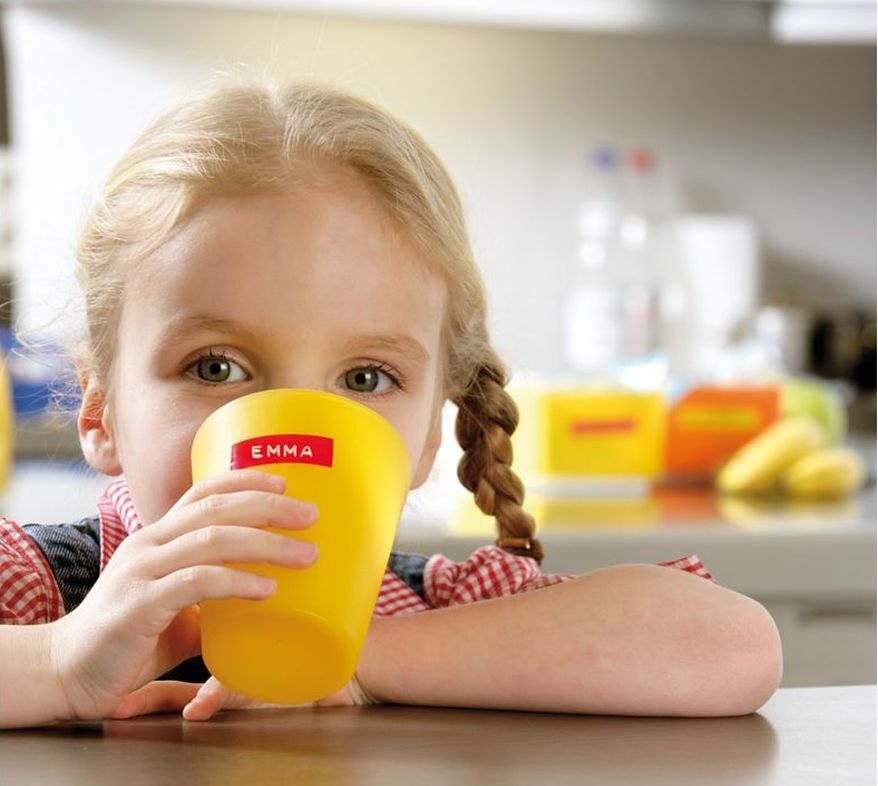 Ein Mädchen lehnt an einer Tischkante und hat einen gelben Plastikbecher in der rechten Hand, aus dem sie etwas trinkt. Der Becher ist mit einem Dymo Prägeband beklebt, auf der ihr Name steht: "Emma".