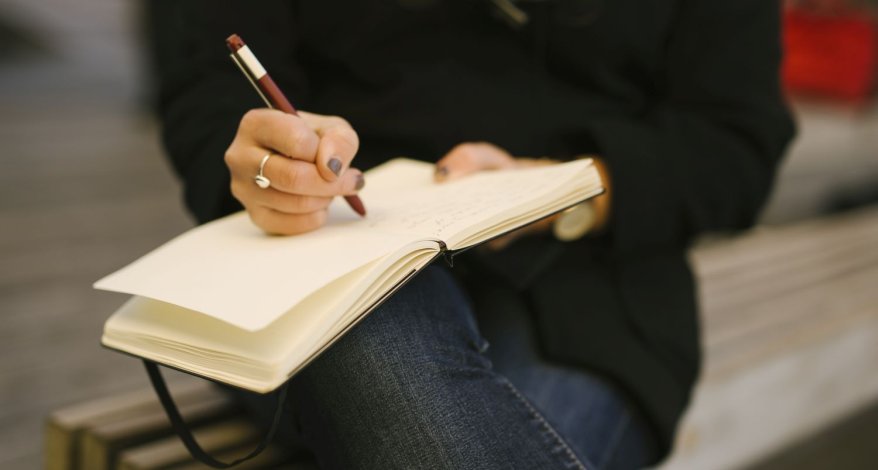 Nahaufnahme einer Frau, die auf einer Bank sitzt und in ein Moleskine Notizbuch schreibt.