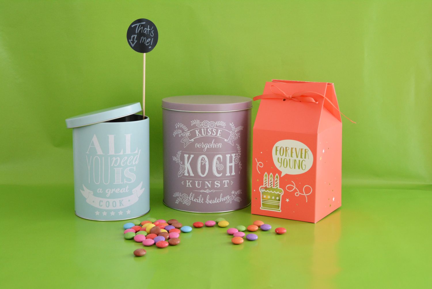 Eine hellblaue und eine lila Metalldose mit Letteringsprüchen sowie eine rote Pappschachtel stehen vor einem grünen Hintergrund. Vor den Dosen und der Schachtel liegen viele bunte Smarties.