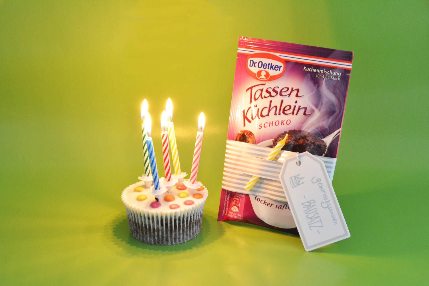 Ein Muffin mit 5 brennenden Kerzen und eine Verpackung zur Zubereitung von Tassen Kuchen stehen vor einem grünen Hintergrund. An der Verpackung hängt ein Etikett der Marke Avery Zweckform mit einer persönlichen Aufschrift.