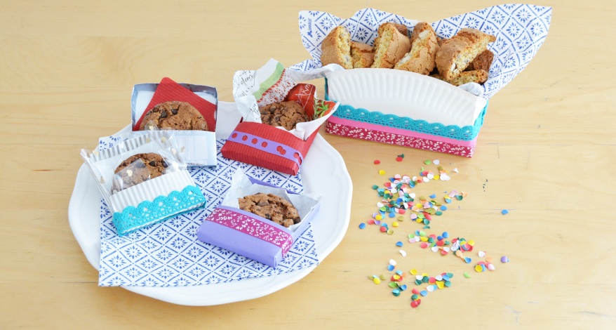 Auf einem Tisch steht ein Teller, auf dem 4 selbstgebastelte Verpackungen für Kekse liegen. Die Verpackungen sind aus buntem Papier und Washi-Tape gebastelt, in den ein Keks liegt. Außerdem steht eine weitere selbstgebastelte Box zur Aufbewahrung von Keksen auf dem Tisch. Die Aufbewahrung ist aus einem Pappteller und buntem Washitape sowie Servietten der Marke Stewo erstellt. 