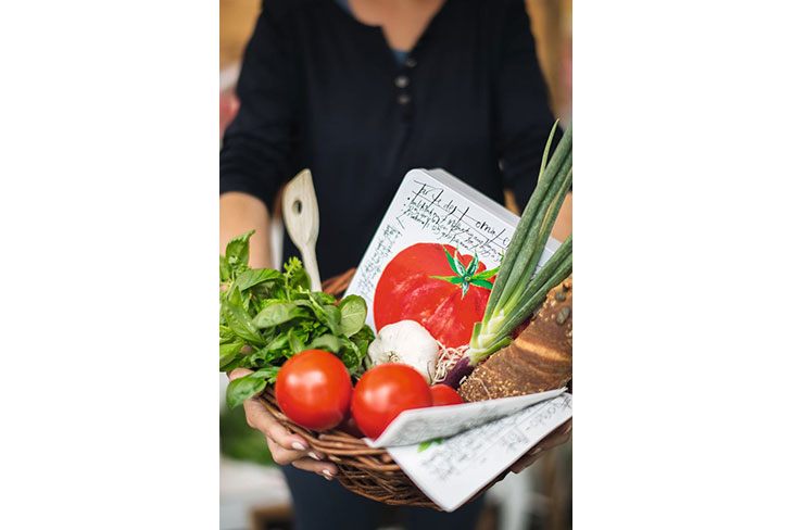 Eine Frau hält einen Korb, der gefüllt mit Tomaten, Knoblauch, Frühlingszwiebeln, Basilikum und einer Metalldose ist, in die Kamera. Die Metalldose ist mit einer Tomate bedruckt.