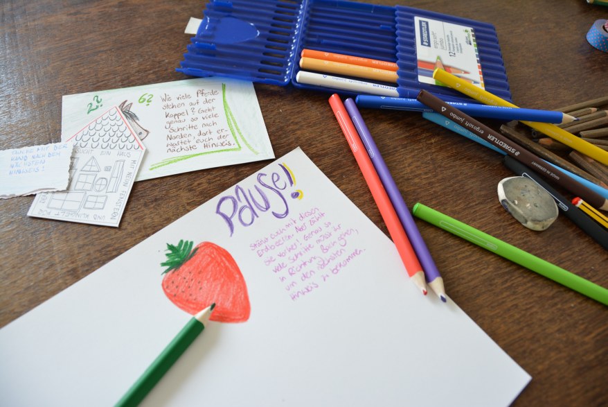 Auf einem Tisch lleigt ein Blatt Papier, auf das mit Buntstifte eine Erdbeere gemalt ist. Daneben steht das Wort "Pause" und ein Text darunter. Auf dem Tisch liegen viele Buntstifte der Marke STAEDTLER sowie die Verpackung der Stifte. 