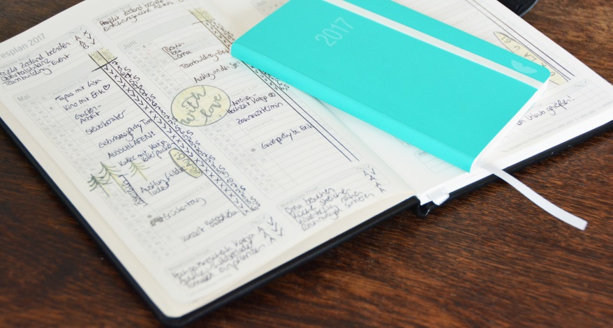 Auf einem Holztisch liegt ein aufgeschlagener Kalender mit vielen Notizen und Einträgen. Darauf liegt ein kleinerer zugeschlagener Kalender.