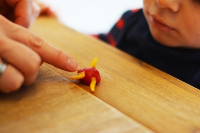 Eine Erwachsenenhand drückt auf den runden Bienenwachs, der auf dem Tisch liegt, um eine Maus zu formen. Das Gesicht einen Kindes, das zuschaut, ist im Anschnitt zu erkennen.