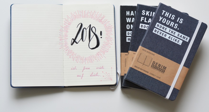 Offenes Notizbuch mit geletterter Zahl "2018" und drei geschlossene Denim-Moleskine-Hefte.