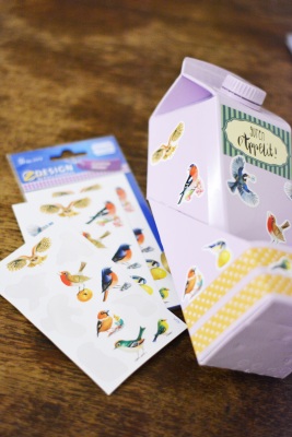 Ein selbstgebasteltes lila Vogelhäuschen, welches aus einem Milchkarton gebastelt wurde, liegt auf einem Holztisch. Es ist mit vierlen bunten Vogel-Stickern beklebt. Daneben liegen Vogelsticker der Marke Avery Zweckform.