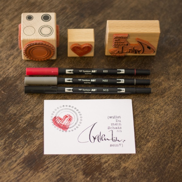 Geletterte Valentinskarte mit Herzmotiv, Stempeln und Tombow-Stiften auf Holztisch.