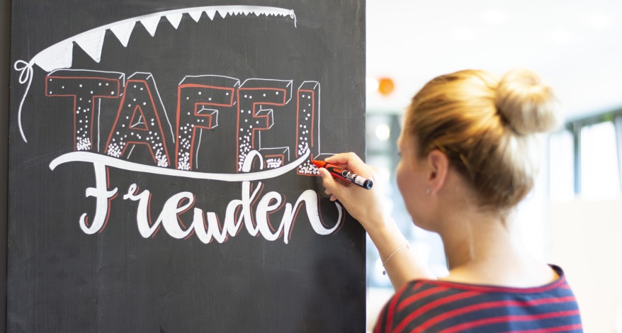 Frau schreibt „Tafel Freuden“ mit Kreidemarkern auf eine schwarze Tafel.