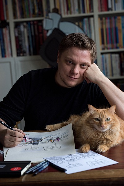 Comiczeichner Michael Holtschulte, der an seinem Schreibtisch sitzt und in die Kamer schaut. Vor ihm liegen Zeichnungen und seine Katze liegt auf dem Tisch neben ihm.