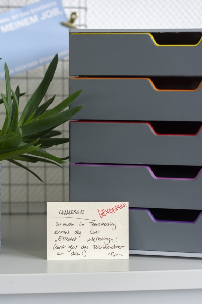 Zettel mit Büro-Challenge neben grauer Schubladenbox und Pflanze