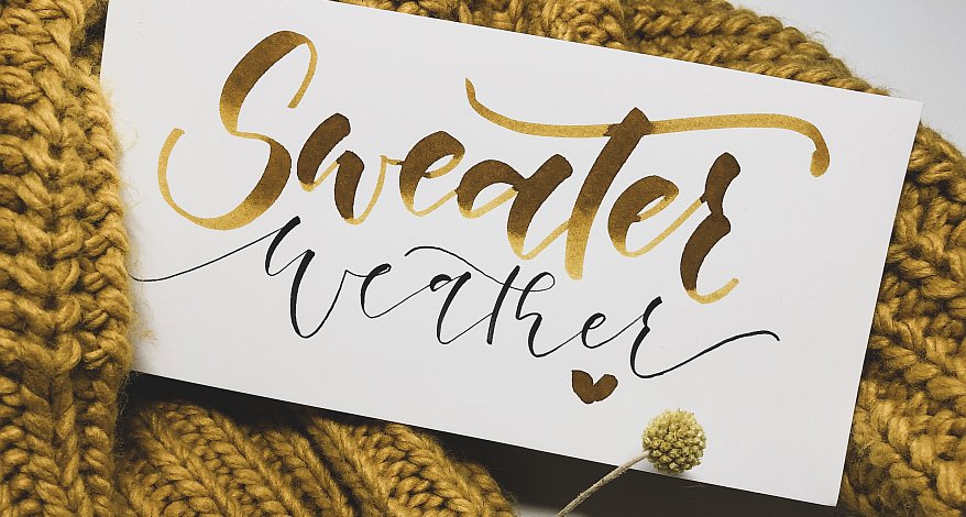Eine Karte mit einem geletterten Spruch "Sweet weather" liegt auf einem okerfarbenem Pullover. 