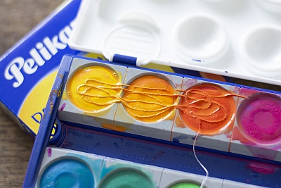 Nahaufnahme eines Pelikan Farbkastens mit den Farben gelb, orange und rot. Darin liegt ein dünner Faden.
