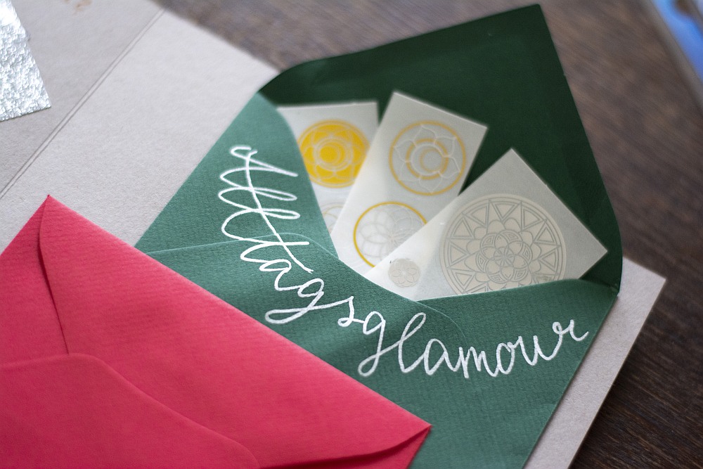 Grüner Umschlag mit Aufschrift „Alltagsglamour“ und Aufklebern