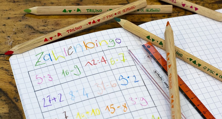 Ein kariertes Heft liegt auf einemHolzuntergrund. Auf dem Heft liegen ein Lineal von Herlitz und verschiedene Buntstifte der Trilino-Produktserie. In das Heft wurde in bunten Farben ein Zahlenbingo mit verschiedenen Rechenaufgaben gemalt.