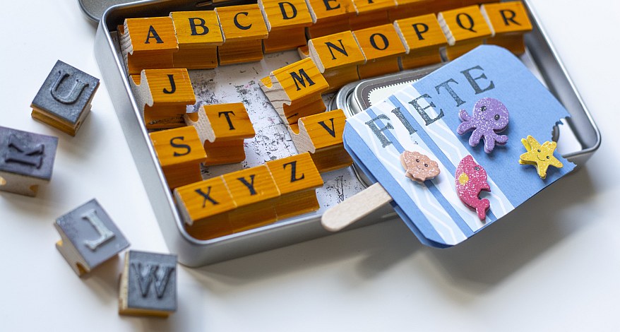 Eine Einladungskarte, die als Eis am Stiel gestaltet ist, liegt auf einem Kasten mit verschiedenen Buchstabenstempeln.