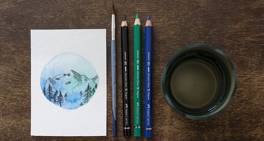 Auf einem Holztisch liegt eine selbstestaltete Karte mit Bergmotiv, Pinsel, Buntstifte und ein Glas