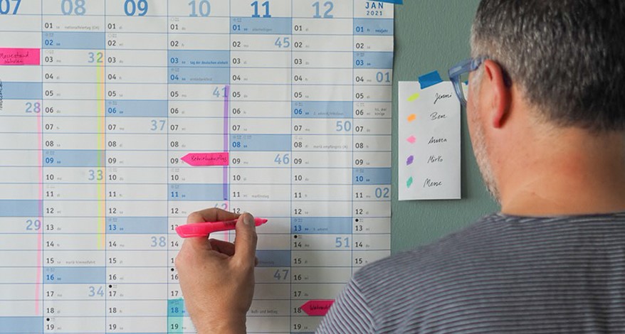 Ein Mann mit kurzen grauen Haaren und einer Brille steht mit dem Rücken zur Kamera. In seiner linken Hand hält er einen pinken Textmarker. Damit markiert er einen Tag in einem Wandkalender, der vor ihm an der Wand hängt.