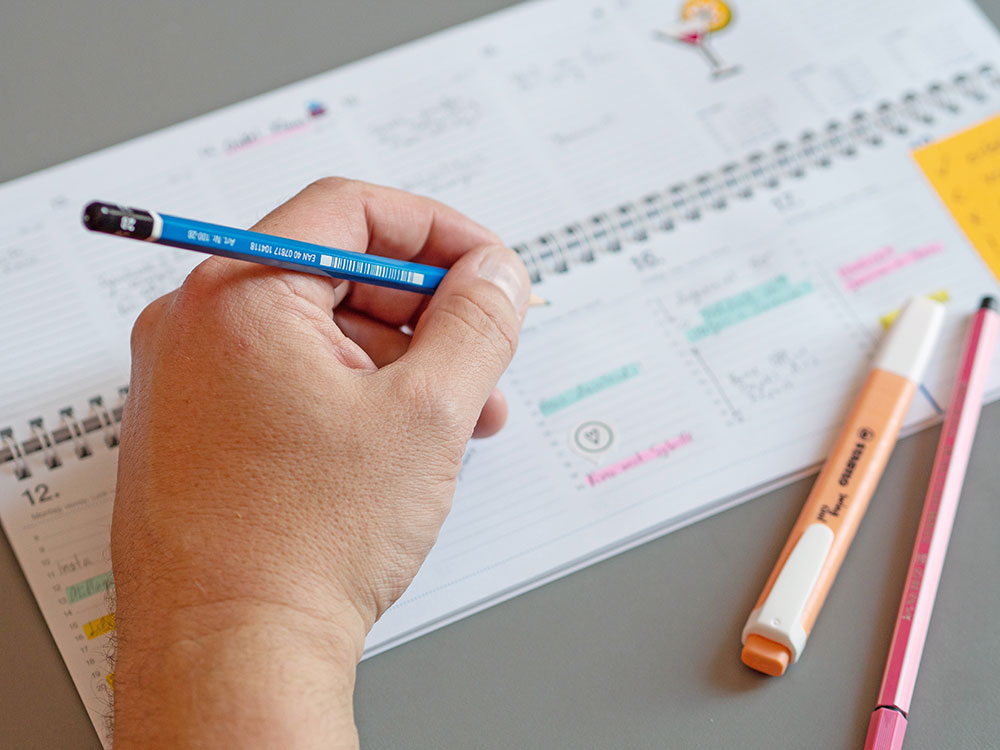 Eine Hand umgreift einen blauen Beistift. Im Hintergrund ist unscharf ein aufgeschlagener Tischkalender der Marke Athesia zu sehen, in den bereits einige Termine eingetragen wurde. Davor liegen ein Textmarker und ein Fasermaler.