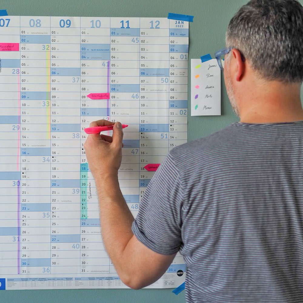 Ein Mann mit kurzen grauen Haaren und einer Brille steht mit dem Rücken zur Kamera. In seiner linken Hand hält er einen pinken Textmarker. Damit markiert er einen Tag in einem Wandkalender, der vor ihm an der Wand hängt.