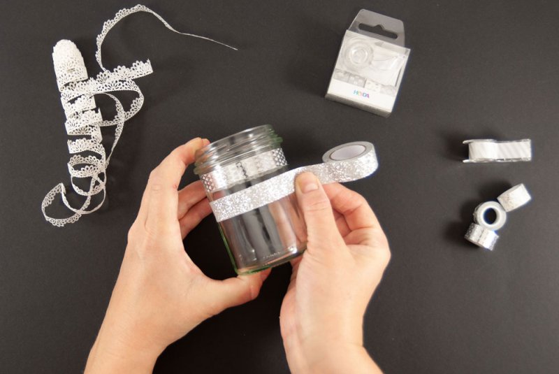 Ein Marmeladenglas wird weiß-grauem mit Washitape der Marke Heyda beklebt. Der Untergrund ist schwarz. Um das Glas verteilt liegen weitere helle Washitapes sowie weiße Washitape-Bordüre. 