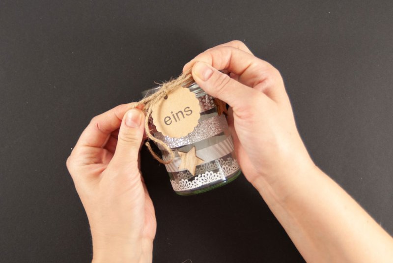 An ein mit weiß-grauem Washitape beklebtes Marmeladenglas, welches mit einem Holzstern der Marke Knorr Prandell beklebt wurde, wird eine braune Kordel befestigt. An dieser hängt ein Geschenkanhänger aus Kraftkarton, auf den das Wort "eins" gestempelt wurde. Der Untergrund ist schwarz.