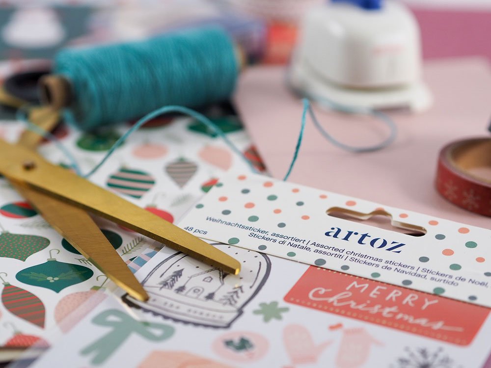Eine goldene Schere liegt auf einer Stickerpackung mit Weihnachtsmotiven der Marke Artoz. Im Hintergrund sind unscharf blauer Garn, ein Washitape, Motivpapiere und ein Motivstanzer zu sehen.