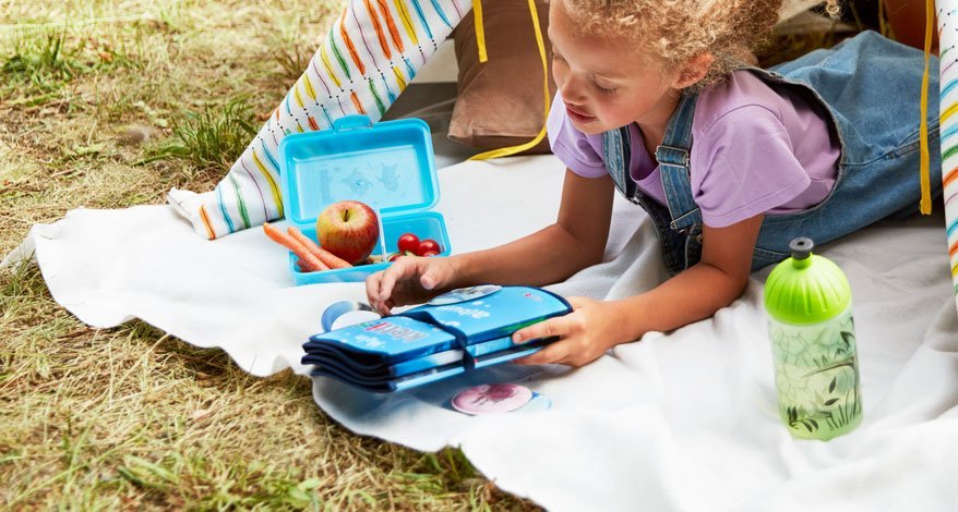 Ein Mädchen liegt im Garten auf einer Decke und hält ein Mäppchen in der Hand. Neben ihm steht eine Trinkflasche und es liegt eine Brotdose mit Obst neben ihm.