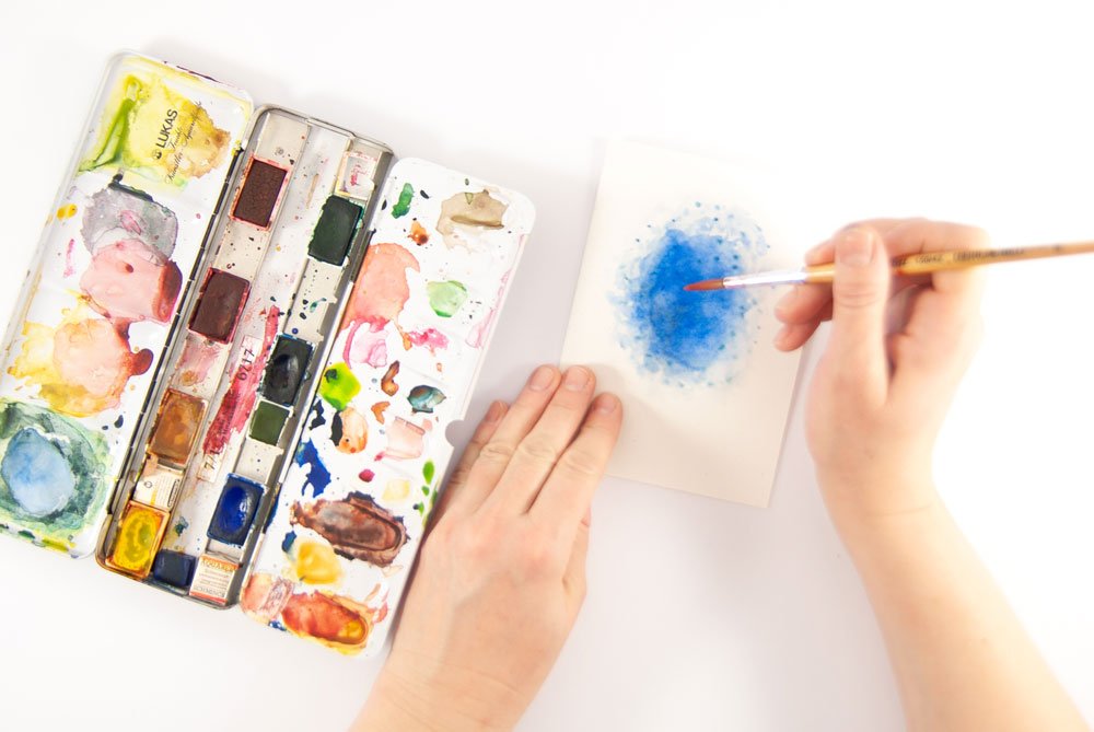 Hände malen mit Aquarellfarben einen blauen Hintergrund auf eine Karte.
