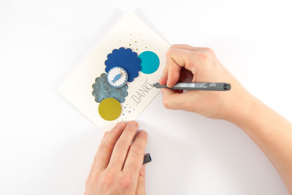 Hand schreibt „DANKE“ auf eine selbst gestaltete Dankeskarte mit bunten Kreisen.