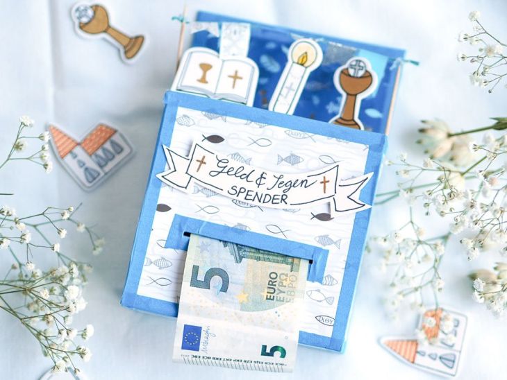 Auf einem weißen Untergrund liegt ein selbstgebasteltes Geschenk zur Kommunion/Konformation. Der "Geld und Segenspender" wurde mit verschiedenen christilichen Symbolen verziert und ein 5€-Schein schaut heraus. Drum herum liegen einige weiße Blumen sowie Sticker.
