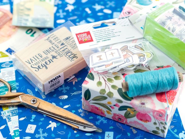 Im Bild sind verschiedenste Bastelmaterialien von Knorr Prandell und Heyda passend zum Thema Kommunio/Konfirmation zu sehen: Verschiedene Motivkartons, ein Stempel, blauer Garn, eine Schere, Washitape, Sticker und eine Geschenkbox. Im oberen Bildrand sind unscharf zwei Geldscheine zu erkennen.
