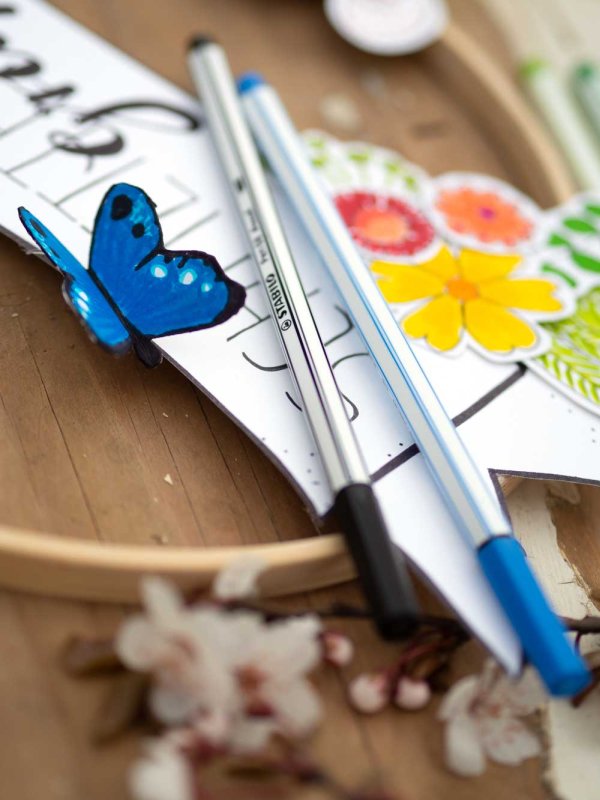 Nahaufnahme: blauer Papier-Schmetterling und Blumen auf Dekoreif, darauf Stabilo Point Stifte