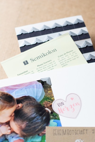 Fotoecken der Marke Semikolon auf einem Blister-Papier, davor sieht man einen Ausdchnitt eines Fotoalbums, wo ein Bild mit einer Fotoecken befestigt ist.