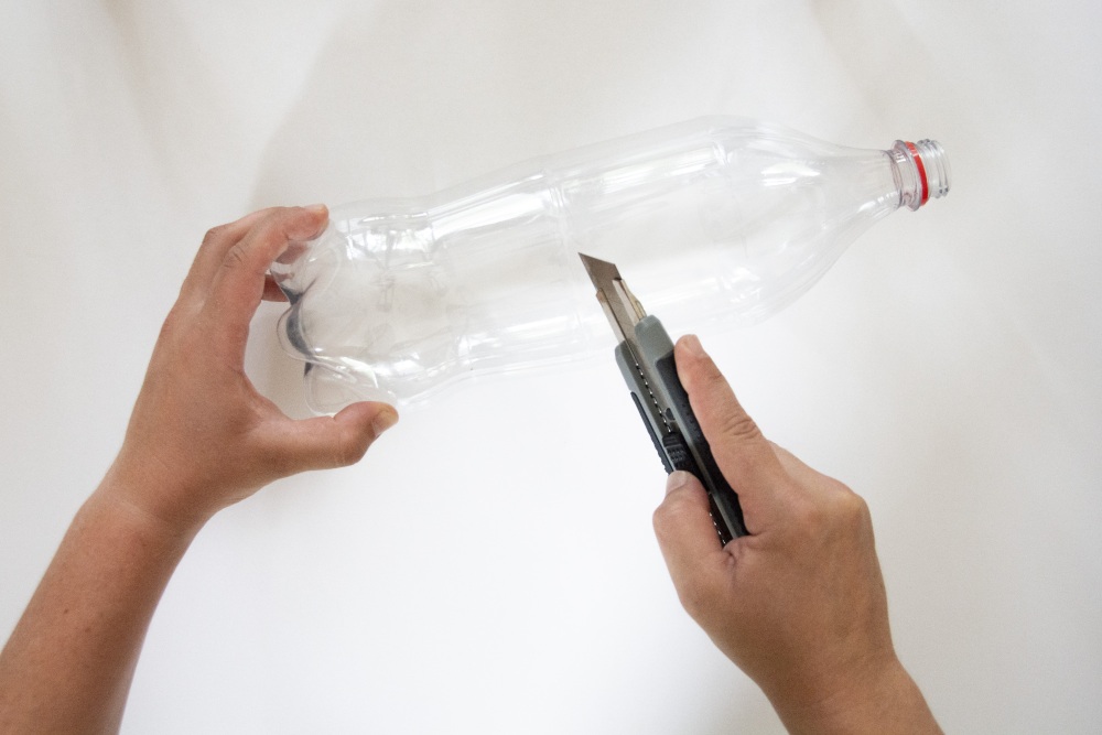 Hände halten eine Plastikflasche, während sie mit einem Cutter-Messer aufgeschnitten wird.