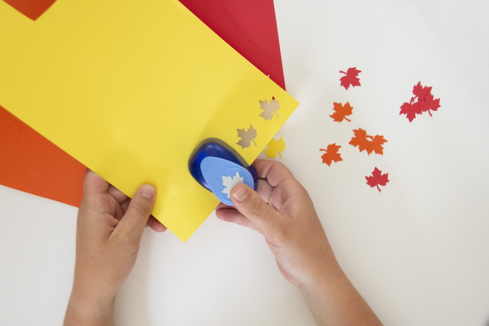 Hände stanzen mit einer Motivstanze bunte Ahornblätter aus gelbem Papier aus.