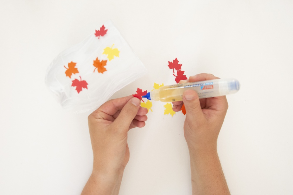 Hände kleben bunte Papier-Ahornblätter mit einem Klebestift auf eine Laterne.