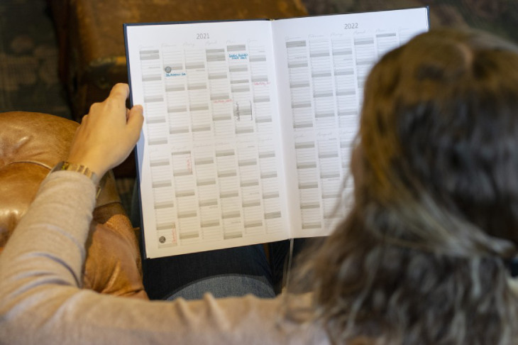 Foto über die Schulter einer jungen Frau, die auf einer Ledercouch sitzt und den aufgeschlagenen 10-Jahreskalender der Marke Brunnen in der Hand hält. 