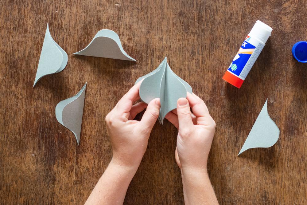 Auf einem Holztisch liegen mehrere Papierelemente und ein Klebestift. Zwei Hände verkleben 2 Papierelemente.