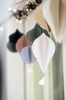 Quasten aus Papier in verschiedenen Formen und Farben hängen an einem Ast mit Lichterkette.