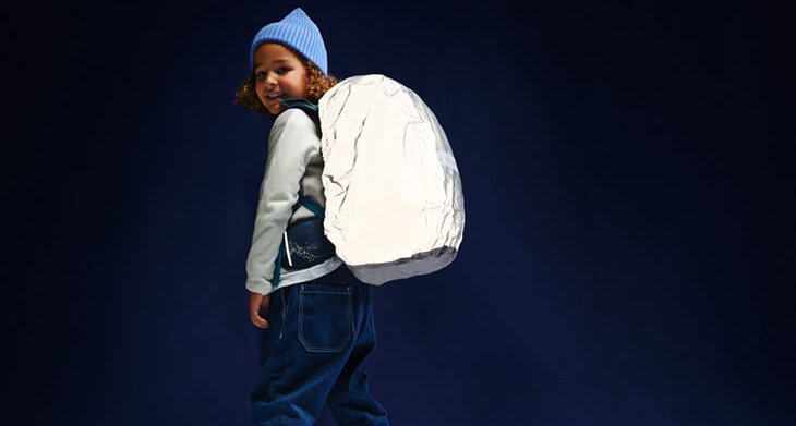 Ein Kind mit Schulranzen und reflektierender Regenhülle von ergobag blickt über die Schulter zur Kamera